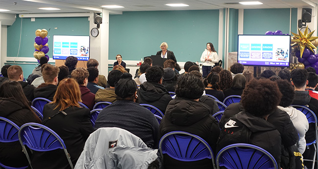 Students attending apprenticeship presentation talks. They are sitting in blue plastic chairs, looking at the speakers in front who have screens either side of them.